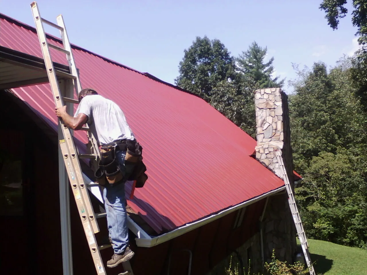 Roofer on a ladder beside a red metal roof and stone chimney — Soffit & Fascia Repair by Roofing Brothers in Breinigsville